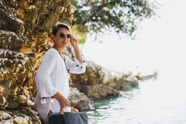 A  woman wearing sunglasses and a white tunic stands by a rocky coastline, holding a laptop bag 