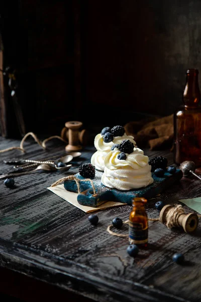 Two homemade white mini desserts pavlova on wooden blue board with whipped cream and blueberries, blackberries on rustic table