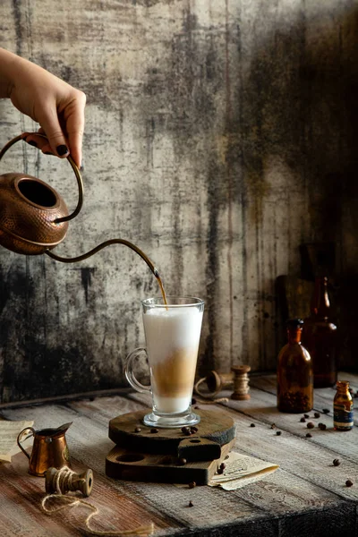 Woman pour coffee from copper kettle in latte glass stands on wooden boards stand on grey rustic table with milk saucer, spool of thread, vintage bottles, beans opposite concrete wall