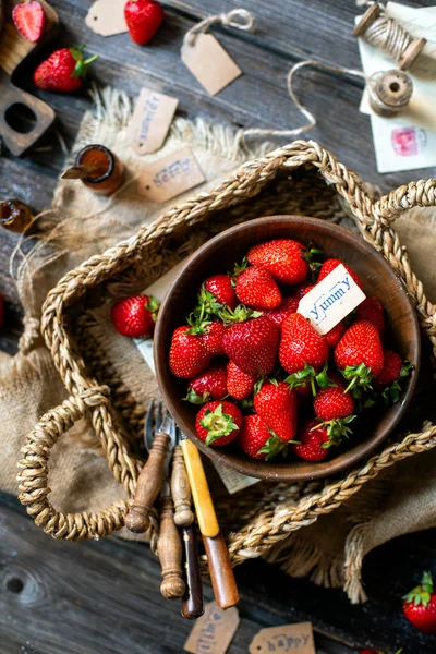 Overhead shot of tasty ripe strawberries in wooden bowl stands in wicker strawy basket on rustic table with sackcloth, bottles, spoons, scissors, bowl with powdered sugar and strainer