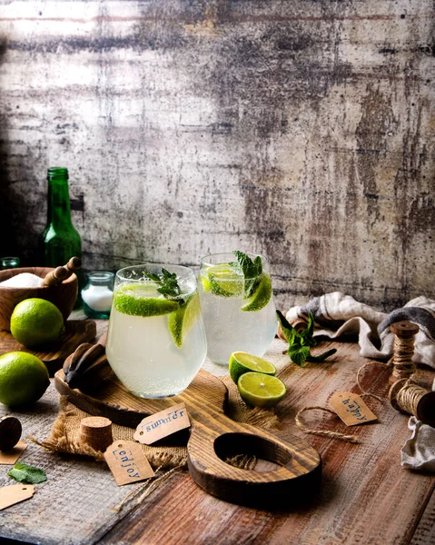 Two glasses of cold icy summer cocktail mojito with mint leaves and slices of lime stands on wooden board on grey table with bowl of sugar, limes, bottles, napkin, tags opposite concrete wall