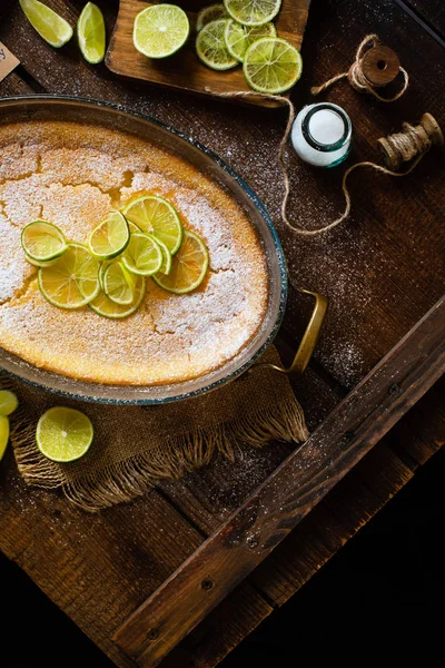 Overhead shot of homemade casserole, pudding, cheesecake, tart, pie or mousse with slices of lime on top in oval glass baking dish stands on rustic wooden brown table with limes, powdered sugar
