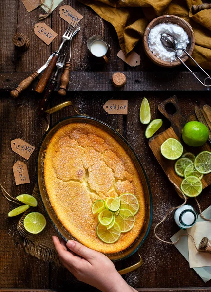 Overhead shot of homemade casserole, pudding, cheesecake, tart, pie or mousse with lime on top in oval baking dish on rustic brown table with limes on board, forks, powdered sugar, strainer, napkin