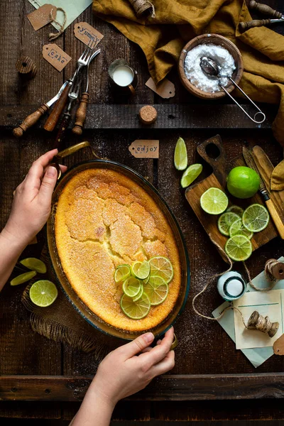 Overhead shot of homemade casserole, pudding, cheesecake, tart, pie or mousse with slices of lime on top in oval baking dish on woman hands on rustic brown table with limes, forks, powdered sugar