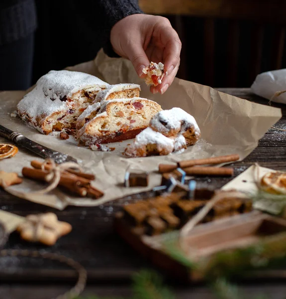 sliced homemade Christmas dessert stollen with dried berries and nuts on parchment in woman hand on wooden rustic table with cinnamon, orange slices, Christmas tree branches, selective focus