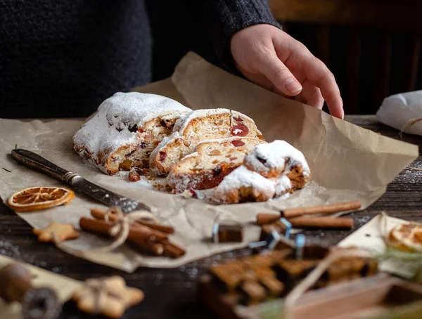 sliced homemade Christmas dessert stollen with dried berries and nuts on parchment in woman hand on wooden rustic table with cinnamon, orange slices, Christmas tree branches, selective focus