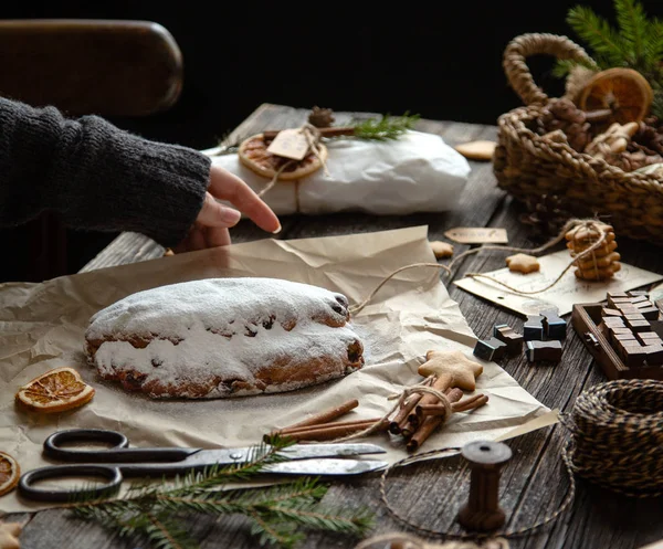 tasty homemade traditional christmas dessert stollen with dried berries and nuts on parchment in woman hand on wooden rustic table with spices, orange slices, Christmas tree branches, selective focus