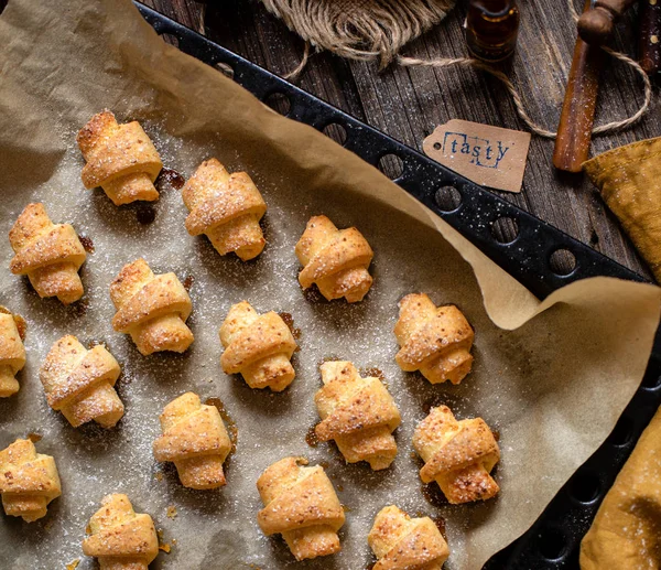 overhead shot of tasty baked rolled or bagels cookies on baking tray with parchment on rustic wooden table with bowls of flour, sugar and cottage cheese, orange napkin