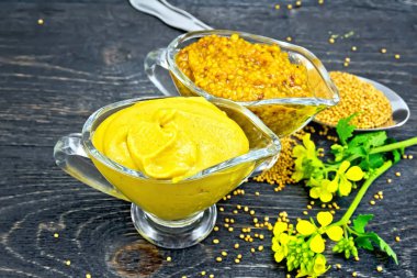 Hardal sosu ve iki cam tencere, sarı çiçek ve tohum içinde ahşap tahta zemin üzerine bir kaşık Dijon hardal