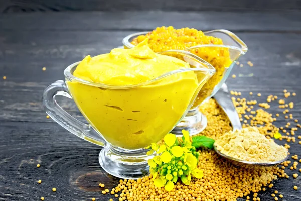 Mustard sauce and Dijon mustard in two glass saucepans, yellow flower, seeds and powder in a spoon on wooden plank background