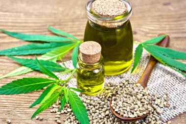 İki cam kavanozda kenevir yağı, çuval bezinde tahıl taneleri olan bir kaşık, eski bir tahtanın arka planında kenevir yaprakları ve sapları