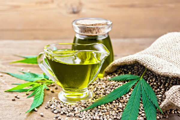 Конопляное масло в стеклянном соусе и банка с зерном в мешке, листья и стебли конопли на старом деревянном фоне доски
