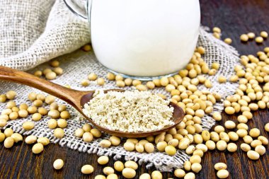 Soy flour in a spoon and milk in a glass jug, soybeans on napkin of burlap on wooden board backgroun
