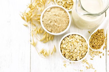 Yulaf gevreği ve un kâselerde, tahıl kaşıkta, yulaf ezmesi sütü cam bir sürahide ve olgun yulaf sapları yukarıdan gelen ahşap bir tahtanın arka planında.