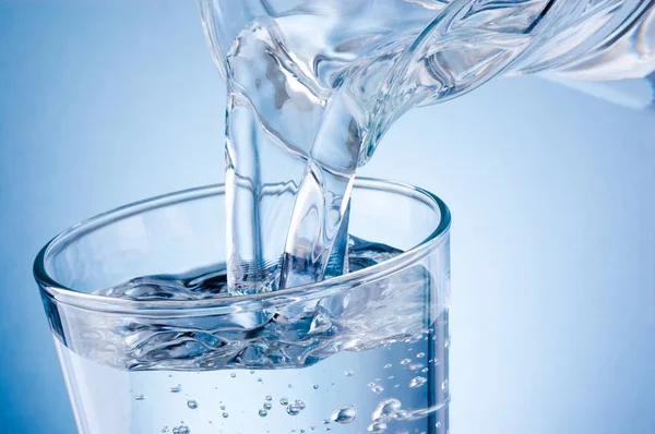 Verter agua de la jarra en un vaso sobre fondo azul — Foto de Stock