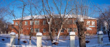 Karlı bir avluda ağaçların arkasında büyük kafes pencereleri ile kırmızı tuğla bina.