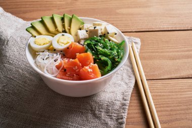kase poke somon, avokado, pirinç, Chuka salata, peynir,, bıldırcın yumurtası serpilir beyaz ve siyah susam ile chopsticks ve soya sosu islated üzerine ahşap arka plan üstten görünüm