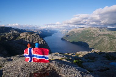 Preikestolen pathway. Couple with the flag of Norway looks at the panorama of the Lysefjord. Tourist attraction. Sunny weather in the mountains clipart