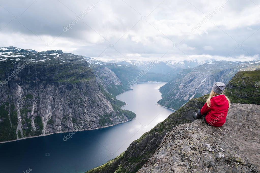 Majestic Trolltunga - one of Norway most spectacular sight. Girl in red jacket sits on rock and looks at Ringedalsvatnet Lake