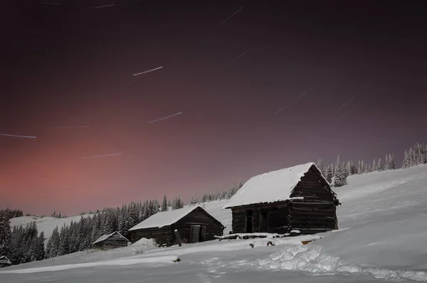 Winter night in a mountain village. Old wooden huts in the snow. Starry sky and moonlight. Long exposure