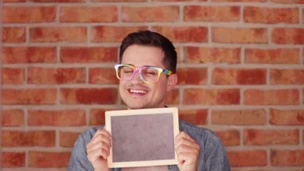 レンガ壁の背景に小さな黒板を保持している眼鏡で幸せな男 — ストック動画