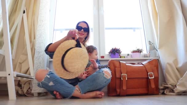 妇女坐在地板与儿子在旅行袋子附近和采取帽子 — 图库视频影像