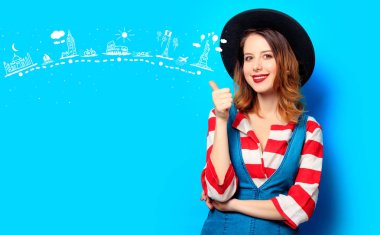 Genç gülümseyen Kızıl saçlı beyaz Avrupalı Bayan kot elbise üzerinde seyahat güzergahı ile mavi arka plan ile şapka ve kırmızı çizgili gömlekli portresi