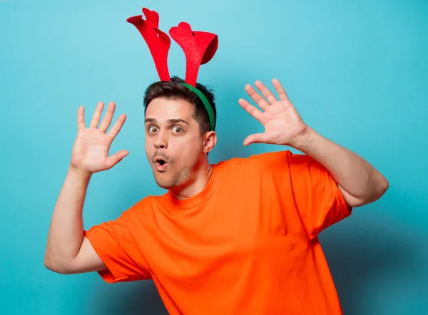 年轻英俊的男人在橙色 T恤与圣诞鹿角 蓝色背景上的工作室图像 — 图库照片