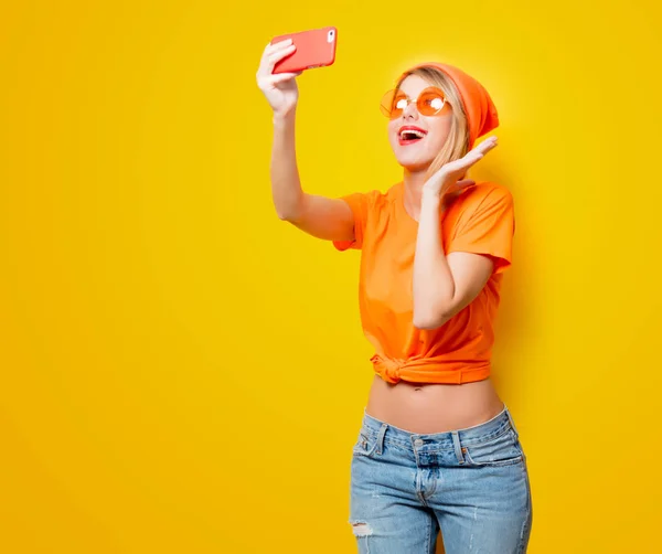 黄色の背景に彼女の携帯電話で Selfie を取ってオレンジ色のメガネを持つ若い女性 1980 年代スタイルの服 — ストック写真