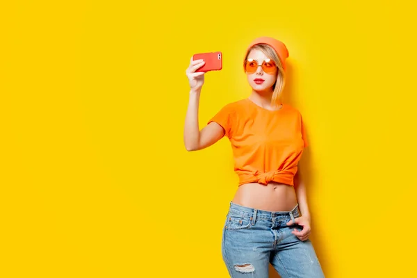 戴着橙色眼镜的年轻妇女带着自拍的手机在黄色背景上 二十世纪八十年代款式服装 — 图库照片
