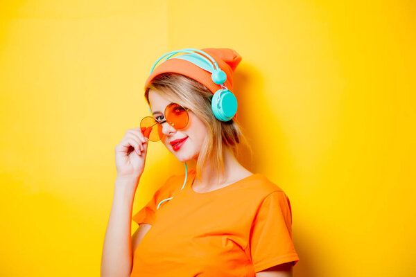 Young stylish woman with orange glasses and headphones on yellow background. Clothes in 1980s style