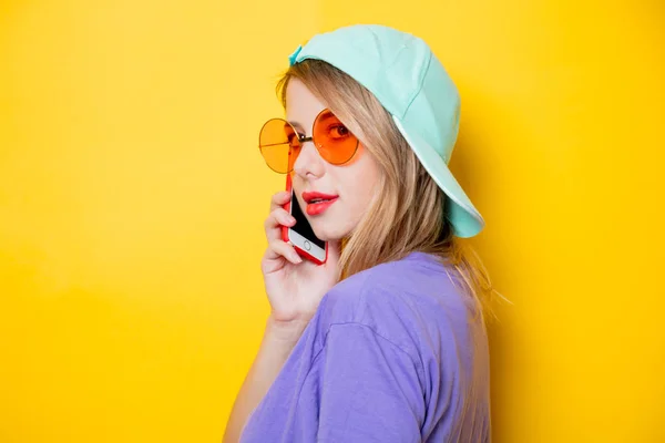 黄色の背景に彼女の携帯電話とオレンジ色のメガネが若いおしゃれな女性 1980 年代スタイルの服 — ストック写真