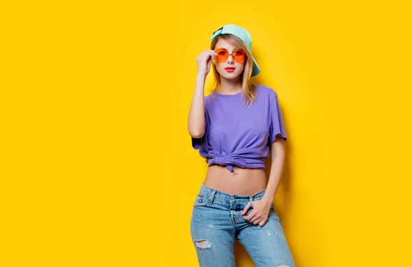 オレンジ色の眼鏡と黄色の背景にキャップを持つ若いスタイルの女の子 1980年代の服装 — ストック写真
