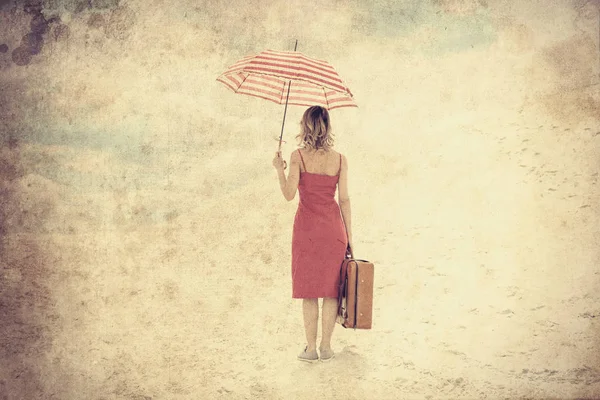 傘とビーチでスーツケースの赤いドレスの若い女性 砂の上旅行コンセプト イメージ 古いカラー画像のスタイルの写真 — ストック写真