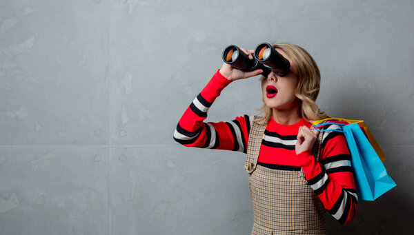 Portrait of a young style blonde girl in red striped sweater with shopping bags and binoculars on grey background