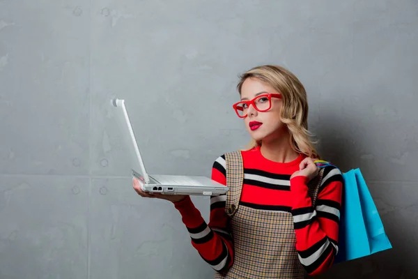 赤いストライプ セーター ショッピング バッグと灰色の背景上のラップトップ コンピューターで若いスタイルのブロンドの女の子の肖像画 — ストック写真