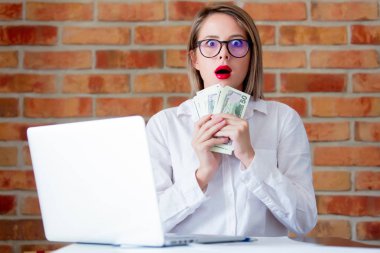 Dizüstü bilgisayar ve tuğla duvar arka plan üzerinde elinde para ile genç iş kadını
