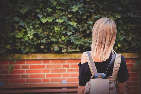 Chica con mochila mirando arbusto y pared de ladrillo —  Fotos de Stock