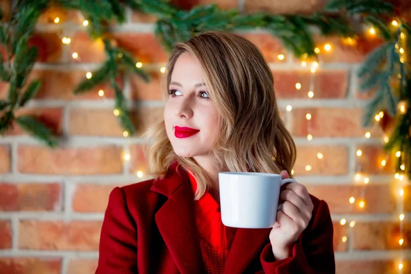 在圣诞节灯光背景下 一个穿着红大衣的年轻女孩的肖像和一杯咖啡 — 图库照片