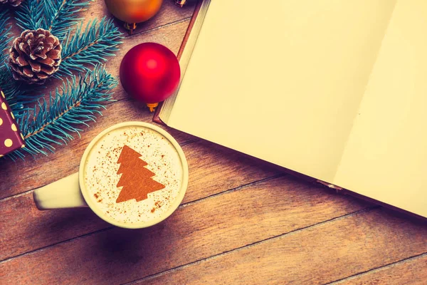 クリームのクリスマス ツリーとおもちゃの近くの木製のテーブルに開いた本カプチーノの白いカップ — ストック写真