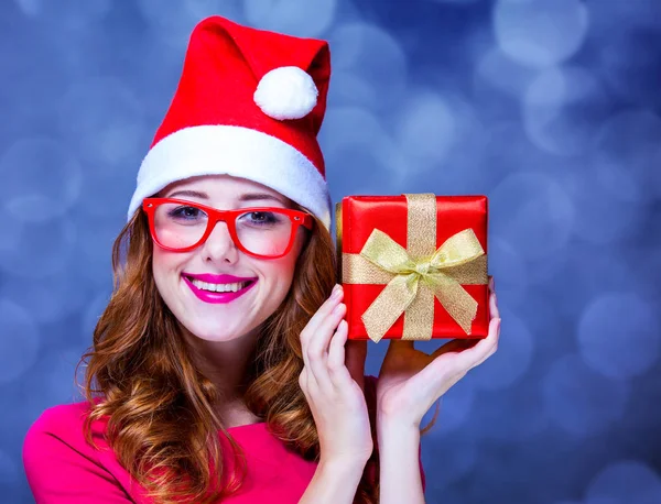 Девушка в рождественской шляпе и очках с подарком на сером фоне — стоковое фото
