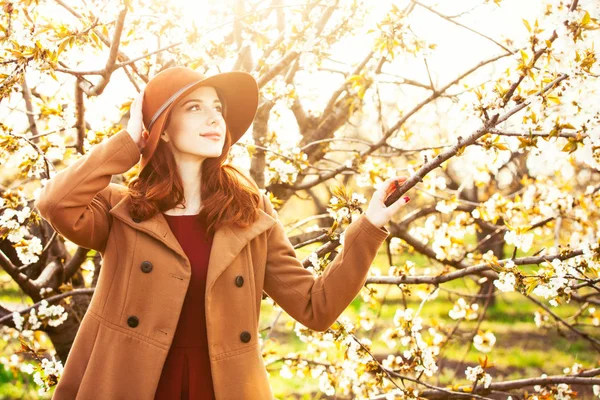 一个美丽的红发女郎的肖像在大衣和帽子在开花苹果树花园在春天的时间日落 — 图库照片