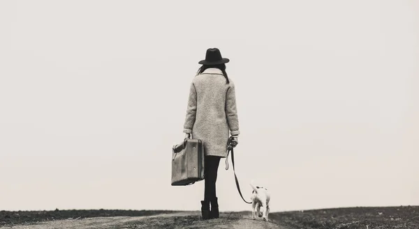 Jovem mulher com mala e cão de pé na estrada — Fotografia de Stock