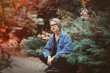 Denim ceket ve gözlük Park zeminde oturan genç kadın. Sonbahar sezonu zaman sahne. Hipster eğilimleri giyim tarzı.