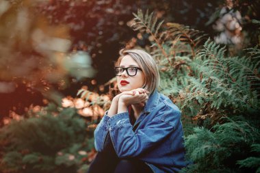 Denim ceket ve gözlük Park zeminde oturan genç kadın. Sonbahar sezonu zaman sahne. Hipster eğilimleri giyim tarzı.