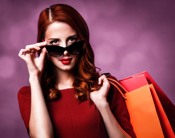 红发女郎在太阳镜与购物袋在紫色背景 — 图库照片