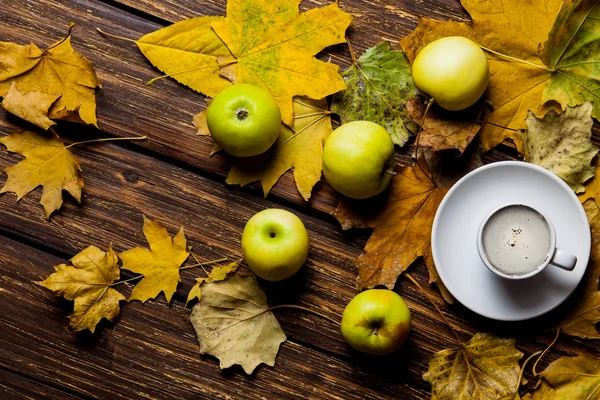 photo of the fallen leaves, apples and white cup of coffee on the brown wooden background