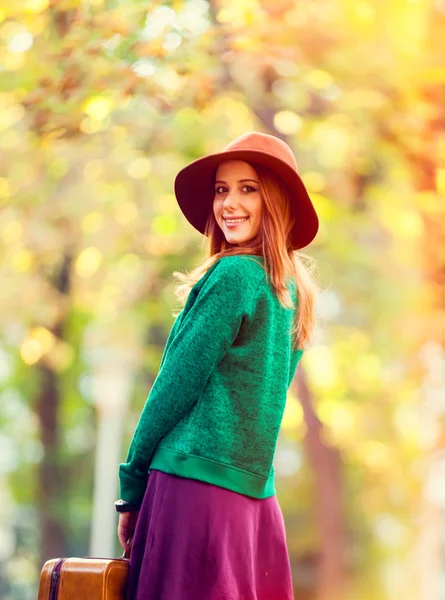 公園でスーツケースと帽子とグリーのセーターで美しい赤毛の女の子 — ストック写真