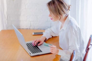 Dizüstü bilgisayar ile bir tablo içinde oturan ve bir kredi kartı tutan beyaz gömlek genç kız. İş, eğitim, alışveriş ve diğer kavramı