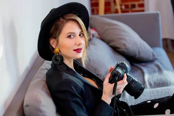 Flicka i svart klänning och hatt med kamera som sitter på en soffa. — Stockfoto
